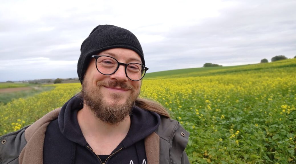 Mann Mitte dreissig, mit spitzzulaufenden Vollbart, kleine Grübchen zieren sein Gesicht. Er trägt einer schwarzen runden Brille einen Bandpulli und steht vor einem Rapsfeld, hinter ihm ist der weite Horizont zu sehen.